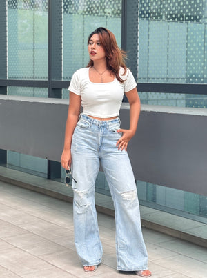A woman modeling a white short-sleeve crop top and light-wash wide-leg distressed jeans, standing outdoors in front of a modern glass building.