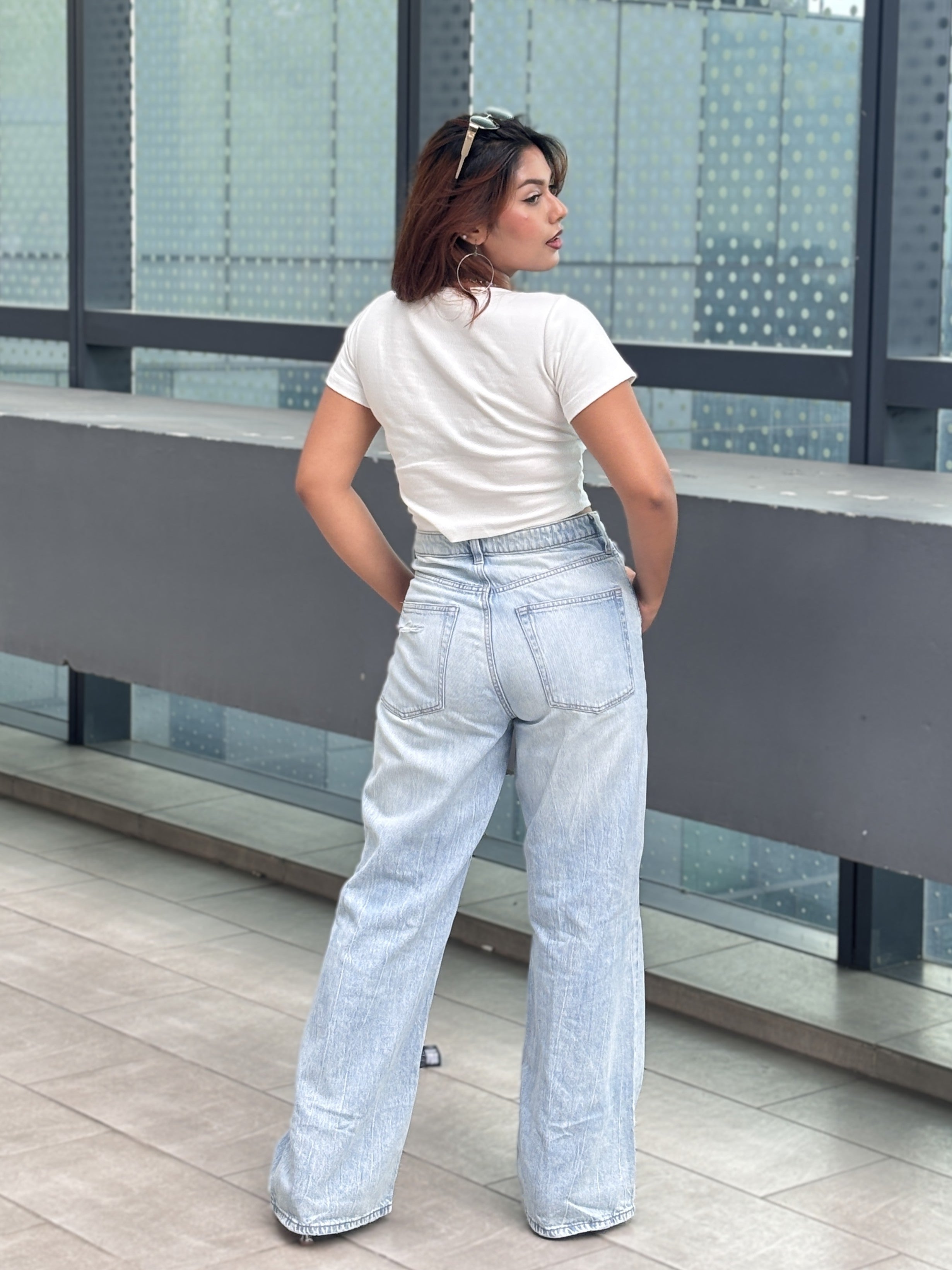 A woman wearing a white t-shirt and light-wash wide-leg jeans, photographed from behind on a modern outdoor balcony with glass windows.