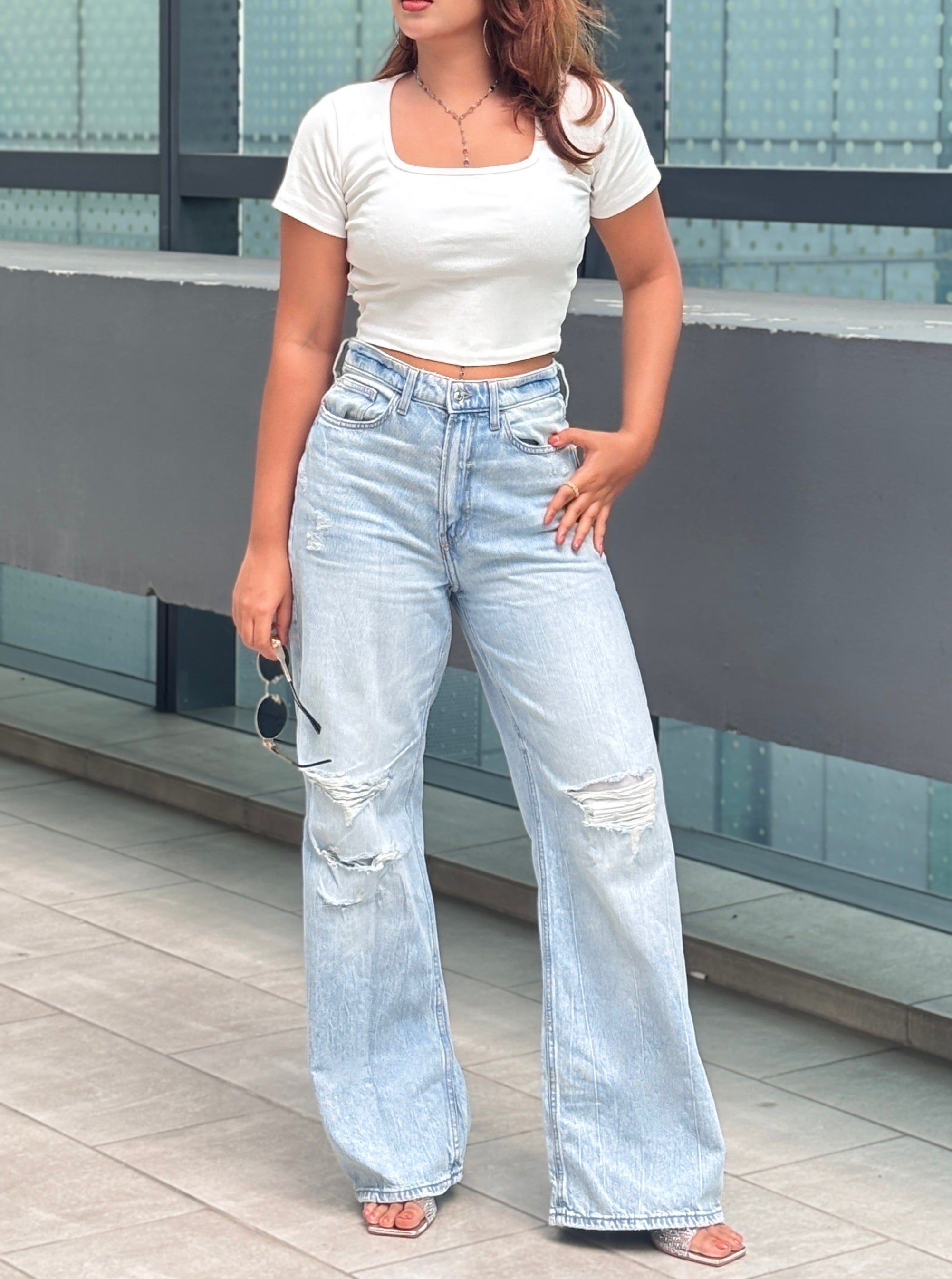 A woman modeling a white short-sleeve crop top and light-wash wide-leg distressed jeans, standing outdoors against a modern glass wall.