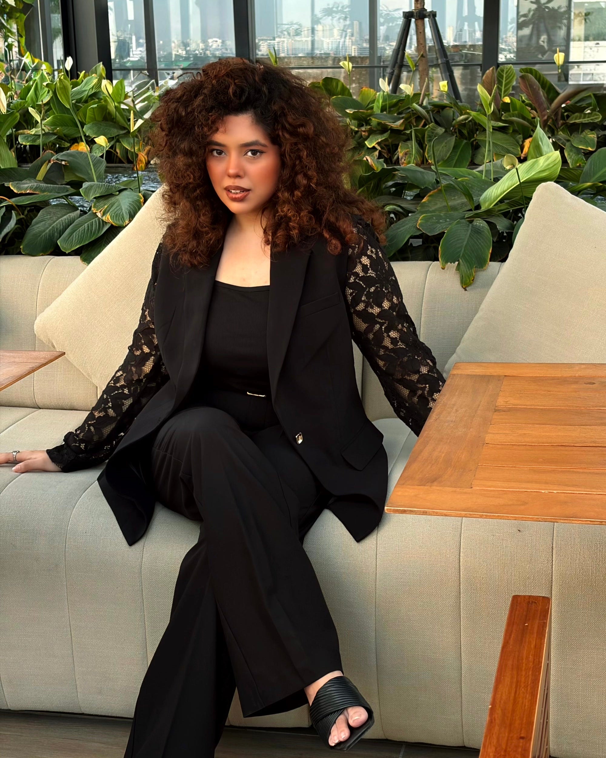 A woman with voluminous curly brown hair sits on a beige outdoor sofa wearing a black lace-trimmed blazer, black top, black trousers, and black slide sandals. The setting features a wooden table in the foreground and lush green plants behind her.