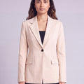 A woman wearing light beige slim-fit formal blazer with notched lapels, front flap pockets, and a single-button closure, styled over a black top and dark trousers. Trendy Buzz BD.